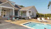 Located at 898–906 Emory Church Rd in Knoxville, TN, this single-level outdoor amenity facility showcases a thoughtfully designed resort-style retreat. The centerpiece is a large swimming pool lined with an extensive row of lounge chairs, offering ample space for sunbathing against a backdrop of mature trees that provide both natural beauty and privacy. Adjacent to the main pool sits a spa/hot tub, seamlessly integrated into the stamped concrete pool deck.

The clubhouse building itself is constructed in a classic craftsman style, featuring board-and-batten siding, stone accent columns, a pitched shingle roof, and white-trimmed multi-pane windows that lend a refined, welcoming aesthetic. A covered porch extends from the building, providing shaded seating directly beside the pool area.

The expansive outdoor patio extends well beyond the pool, offering a generous gathering space complete with wicker lounge furniture, cornhole boards, and a covered pergola housing an outdoor grill — ideal for social events and recreation. Shaded umbrella tables provide additional seating for al fresco dining. Black metal railings frame the various outdoor zones, maintaining a cohesive and polished look throughout. The entire space is enveloped by lush landscaping and open sky, creating a bright, airy, and inviting community atmosphere.