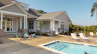 Located at 898–906 Emory Church Rd in Knoxville, TN, this single-level outdoor amenity facility showcases a thoughtfully designed resort-style retreat. The centerpiece is a large swimming pool lined with an extensive row of lounge chairs, offering ample space for sunbathing against a backdrop of mature trees that provide both natural beauty and privacy. Adjacent to the main pool sits a spa/hot tub, seamlessly integrated into the stamped concrete pool deck.

The clubhouse building itself is constructed in a classic craftsman style, featuring board-and-batten siding, stone accent columns, a pitched shingle roof, and white-trimmed multi-pane windows that lend a refined, welcoming aesthetic. A covered porch extends from the building, providing shaded seating directly beside the pool area.

The expansive outdoor patio extends well beyond the pool, offering a generous gathering space complete with wicker lounge furniture, cornhole boards, and a covered pergola housing an outdoor grill — ideal for social events and recreation. Shaded umbrella tables provide additional seating for al fresco dining. Black metal railings frame the various outdoor zones, maintaining a cohesive and polished look throughout. The entire space is enveloped by lush landscaping and open sky, creating a bright, airy, and inviting community atmosphere.