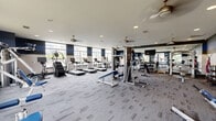 Located at 7145 Anderson Dr in Whitestown, Indiana, this fitness center offers a spacious, single-floor layout designed to support a comprehensive workout experience. The gym is generously proportioned, providing ample room for both cardio and strength training activities without feeling crowded.

The cardio zone features a row of treadmills and elliptical trainers positioned along large, multi-pane windows that flood the space with natural light and provide views of the surrounding neighborhood. Ceiling fans are distributed throughout, contributing to a comfortable and well-ventilated environment.

The strength training area is well-appointed with a multi-station cable machine, various selectorized strength machines, and a dedicated free weight section complete with a full dumbbell rack and adjustable benches. A large wall mirror enhances the sense of space and allows members to monitor their form during workouts. Additional accessories such as resistance bands, stability balls, and a TRX suspension system round out the functional training options.

The interior design features a modern color scheme of navy blue, gray, and white, with patterned carpet tile flooring throughout most of the space and rubber mat flooring in the free weight area. Recessed lighting complements the natural light from the windows. The overall ambiance is clean, functional, and motivating — well-suited for a wide range of fitness routines.
