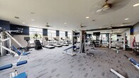 Located at 7145 Anderson Dr in Whitestown, Indiana, this fitness center offers a spacious, single-floor layout designed to support a comprehensive workout experience. The gym is generously proportioned, providing ample room for both cardio and strength training activities without feeling crowded.

The cardio zone features a row of treadmills and elliptical trainers positioned along large, multi-pane windows that flood the space with natural light and provide views of the surrounding neighborhood. Ceiling fans are distributed throughout, contributing to a comfortable and well-ventilated environment.

The strength training area is well-appointed with a multi-station cable machine, various selectorized strength machines, and a dedicated free weight section complete with a full dumbbell rack and adjustable benches. A large wall mirror enhances the sense of space and allows members to monitor their form during workouts. Additional accessories such as resistance bands, stability balls, and a TRX suspension system round out the functional training options.

The interior design features a modern color scheme of navy blue, gray, and white, with patterned carpet tile flooring throughout most of the space and rubber mat flooring in the free weight area. Recessed lighting complements the natural light from the windows. The overall ambiance is clean, functional, and motivating — well-suited for a wide range of fitness routines.
