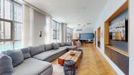 This single-floor space is designed as a multifunctional amenity suite, combining social, work, and leisure areas within a cohesive, modern interior. Throughout, light-toned herringbone hardwood flooring creates a warm visual thread connecting each zone.

The expansive open-plan lounge anchors the space, featuring a generous sectional sofa, a rustic wood coffee table, and floor-to-ceiling windows dressed in sheer white curtains that flood the area with natural light and offer views of the surrounding urban streetscape. The lounge flows seamlessly into a dining area distinguished by a bold blue accent wall, coordinating banquette seating, and a sleek chandelier overhead.

Adjacent, a game room provides a long communal table, a shuffleboard table, and large glass doors opening directly to an outdoor terrace. A co-working area nearby offers long wooden worktables, mixed seating, and additional access to the outdoors.

A well-appointed kitchen showcases dark matte cabinetry, a metallic backsplash, stone countertops, and a central island. A dedicated beverage station with warm wood paneling adds convenience. A lounge reception area with low-profile seating, curated artwork, and natural wood double doors completes the space, contributing to its refined, hospitality-inspired ambiance.