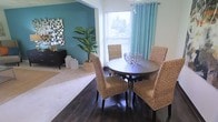 This single-story space showcases an open-concept layout that seamlessly connects the dining and living areas, creating a welcoming and cohesive flow throughout. The dining area features rich, dark hardwood flooring that grounds the space, complemented by a round dark-wood table surrounded by woven rattan chairs that introduce natural texture and a casual, coastal aesthetic. A decorative bowl centerpiece adds an elegant finishing touch to the dining setup. A bright window dressed with teal curtains allows natural light to filter in, offering a view of lush greenery outside. Adjacent to the dining area, the living space transitions to plush carpeting, providing a soft contrast underfoot. A bold teal accent wall serves as a striking focal point, adorned with a mosaic-style mirror arrangement that adds visual depth and interest. A dark wood credenza and a large tropical potted plant further enhance the area's relaxed yet curated atmosphere. The overall ambiance of the space is bright, airy, and thoughtfully styled, blending earthy natural materials with vibrant pops of teal to create an inviting and cohesive interior environment.