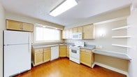 This single-floor space offers a practical and comfortable layout designed for everyday living. The kitchen is a standout feature, showcasing light maple cabinetry that provides generous storage space along both the upper and lower walls. Grey laminate countertops extend across the full length of the kitchen, offering ample workspace for meal preparation. White appliances, including an electric range, built-in microwave, and dishwasher, are seamlessly integrated into the cabinetry for a clean, cohesive look. A stainless steel sink is positioned beneath a large window that allows natural light to fill the space, creating a bright and inviting atmosphere. The warm hardwood-style flooring runs throughout, adding a touch of warmth and continuity to the interior. Overhead, a large flush-mount fluorescent light fixture ensures the kitchen is well-lit at all times. A built-in desk or breakfast bar area is tucked along one wall, providing a convenient spot for casual dining or work. The neutral wall tones complement the light cabinetry and flooring, contributing to an overall clean and airy ambiance throughout the space.