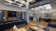 This single-floor space showcases a thoughtfully designed open-plan layout that seamlessly integrates multiple functional zones. The interior immediately draws attention to its dramatic coffered ceiling, detailed with dark wood beams and linear pendant lighting accented by brass hardware, creating a sophisticated architectural focal point overhead.

The main lounge area is furnished with contemporary tan upholstered seating arranged around a low oval coffee table, offering a comfortable and inviting gathering space. A sleek console table lined with lush orchid arrangements adds a refined, decorative touch along one wall.

Toward the rear, a well-appointed kitchenette features warm wood cabinetry, stainless steel appliances, a range hood, and a central island with counter seating, making it well-suited for communal use. Adjacent to the kitchen, a dining area with dark chairs provides additional seating options.

Polished hardwood flooring runs throughout, reflecting the recessed and pendant lighting and enhancing the warmth of the space. Frosted glass-paneled private rooms or office spaces are visible along one wall, offering separation without sacrificing the open feel. The overall ambiance is modern, professional, and welcoming.