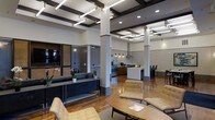 This single-floor space showcases a thoughtfully designed open-plan layout that seamlessly integrates multiple functional zones. The interior immediately draws attention to its dramatic coffered ceiling, detailed with dark wood beams and linear pendant lighting accented by brass hardware, creating a sophisticated architectural focal point overhead.

The main lounge area is furnished with contemporary tan upholstered seating arranged around a low oval coffee table, offering a comfortable and inviting gathering space. A sleek console table lined with lush orchid arrangements adds a refined, decorative touch along one wall.

Toward the rear, a well-appointed kitchenette features warm wood cabinetry, stainless steel appliances, a range hood, and a central island with counter seating, making it well-suited for communal use. Adjacent to the kitchen, a dining area with dark chairs provides additional seating options.

Polished hardwood flooring runs throughout, reflecting the recessed and pendant lighting and enhancing the warmth of the space. Frosted glass-paneled private rooms or office spaces are visible along one wall, offering separation without sacrificing the open feel. The overall ambiance is modern, professional, and welcoming.