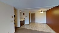 This single-floor residential space is designed with an open-concept layout that seamlessly connects the kitchen and living areas. The kitchen features dark wood cabinetry, a light-toned countertop that doubles as a breakfast bar, and white appliances, including a refrigerator. A half-wall partition separates the kitchen from the adjacent living space, creating a sense of definition without sacrificing openness. The entryway is finished with light-toned tile flooring, which transitions into plush carpeting throughout the living area, offering a warm and comfortable underfoot experience. A ceiling fan with an integrated light fixture is centrally positioned in the living area, providing both air circulation and ambient lighting. The front entry door is finished in a natural wood tone, complementing the cabinetry throughout. Walls are painted in a neutral, light beige tone, contributing to a bright and welcoming atmosphere. The textured ceiling adds a classic residential character to the space. Overall, the layout is practical and efficient, making good use of the available floor plan while maintaining a cohesive and functional design throughout.