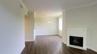 This single-story space showcases a thoughtfully designed open layout that seamlessly connects the living and dining areas. The interior is finished with rich, dark hardwood flooring that contrasts beautifully against the crisp white walls and bright white baseboards, creating a clean and polished aesthetic throughout.

Crown molding lines the ceilings, adding a touch of classic elegance to the space. A distinctive arched entryway on one side provides architectural character and frames the transition between rooms. The dining area is anchored by a delicate chandelier, lending a refined focal point to the space, while a window allows natural light to filter in, brightening the area.

On the opposite side of the room, a white-painted fireplace with a sleek black firebox serves as a standout architectural feature, offering both visual interest and a cozy centerpiece for the living area. A half-wall with a pass-through opening connects to an adjacent room, enhancing the open feel while maintaining a degree of separation between spaces.

Overall, the space presents a neutral, light-filled palette with classic detailing that provides a versatile foundation adaptable to a variety of interior styles.