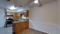 This single-story space showcases a practical and straightforward layout, with the kitchen serving as a central feature. The kitchen is equipped with warm-toned oak cabinetry, offering ample upper and lower storage. Laminate countertops provide functional workspace, and the sink is positioned along the counter with standard fixtures. A peninsula-style counter extension creates a natural transition between the kitchen and the adjacent carpeted area, which could serve as a dining or living space.

The flooring in the open adjacent area is carpeted in a neutral tone, contributing to a cozy atmosphere. White walls with a classic chair rail detail run along the perimeter, adding a subtle architectural touch. The ceiling features a basic light fixture with a pull-chain design, providing straightforward overhead lighting. A fluorescent light above the kitchen workspace ensures adequate task lighting.

Overall, the space presents a clean, utilitarian design with a neutral color palette. The open connection between the kitchen and the adjacent area promotes a sense of flow, making the layout practical for everyday living.