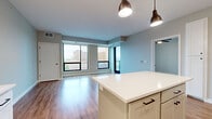 This modern single-floor residence offers a thoughtfully designed open-concept layout that seamlessly connects the main living areas. The kitchen is a standout feature, equipped with stainless steel appliances including a French door refrigerator, gas range, microwave, and dishwasher. Crisp white shaker cabinetry, a light subway tile backsplash, quartz countertops, and a central island with pendant lighting complete the space, which flows directly into the living area. The living room is bright and airy, benefiting from floor-to-ceiling windows framed in sleek black that offer sweeping urban views and flood the space with natural light. Hardwood-style flooring runs throughout the main living areas and hallways, providing a warm and cohesive aesthetic. The bedrooms are carpeted for comfort, each featuring large windows with city views and ceiling fans where applicable. The bathroom is finished with wood-look flooring, a walk-in shower enclosed by a black-framed glass door, and clean white tile work. A well-proportioned hallway connects the rooms, maintaining an efficient and functional flow throughout the unit. The overall palette of soft grays, warm neutrals, and white trim lends the space a clean, contemporary character.