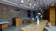 The Shop — Workers Union is a single-floor coworking and communal space designed with a distinct industrial aesthetic. The interior features exposed brick walls adorned with hand-lettered signage, including the prominent branding of "The Shop" and an "Internet Ready" declaration, reinforcing the space's identity as a connected workspace. A striking pixel-art mural anchors one end of the room, adding a creative and contemporary focal point.

The ceiling is open and utilitarian, with exposed conduit, ventilation ducts, and a cascading arrangement of Edison bulb pendant lights strung from a track system, casting a warm, ambient glow throughout the space. Large communal wooden tables provide generous shared work surfaces, while custom wood-framed booth seating with plaid upholstery offers a more intimate setting for focused work or small group meetings.

The flooring is a smooth concrete in a muted gray tone, complementing the raw, urban character of the overall design. Decorative touches such as white floral arrangements in tall vases soften the industrial palette. The space balances a rugged, creative atmosphere with functional, well-considered furnishings, making it well-suited for professionals, freelancers, and collaborative teams seeking an inspiring and connected work environment.