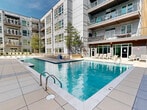 Located in the heart of Charlotte's Brooklyn Village neighborhood, this outdoor amenity deck offers a resort-inspired retreat within an urban setting. The centerpiece is a generously sized swimming pool with crystal-clear water, framed by wide, light-toned tile surrounds that provide ample space for lounging. Rows of modern chaise loungers are thoughtfully arranged poolside and on adjacent turf areas, accommodating both sunbathing and relaxed socializing.

A dedicated spa section features built-in water jet loungers, creating a hydrotherapy experience alongside the main pool. Nearby, a stylishly appointed outdoor bar and lounge area is anchored by string lights overhead and surrounded by wicker seating, ideal for gathering in the evening. Mature trees and curated landscaping add natural greenery throughout the courtyard, softening the contemporary architectural backdrop of the multi-story building with its distinctive mix of glass, steel, and warm wood-toned paneling.

The deck also includes a pergola-covered retreat and open-air seating areas that frame views of the Charlotte city skyline. The combination of modern design, thoughtful amenities, and an open, sunlit atmosphere makes this outdoor space a standout feature of this urban property.