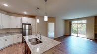This single-floor residence offers a thoughtfully designed open-concept layout that seamlessly connects the kitchen, dining, and living areas. The kitchen serves as the centerpiece, featuring crisp white shaker cabinetry, light granite countertops, and a large island with a double stainless steel sink illuminated by stylish pendant lights. Stainless steel appliances, including a refrigerator, range, and microwave, complete the modern culinary space. Warm hardwood-style flooring flows throughout the main living areas, while a sliding glass door in the living room floods the space with natural light and provides outdoor access. Built-in shelving adds functional character to the living area. The bedrooms are carpeted and bright, each featuring windows with lush wooded views; one includes a ceiling fan for added comfort. Two bathrooms serve the residence — one with a soaking tub and shower combination alongside a dark wood vanity with white countertop, and another with direct access to a spacious walk-in closet outfitted with built-in shelving. Throughout, neutral warm-toned walls create a cohesive and inviting atmosphere across every room.