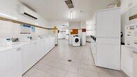 Located at 80 Forest Manor Rd in Toronto, this shared laundry facility offers a generously sized, single-floor space designed for efficient, high-capacity use. The room is bright and clean, featuring white ceramic tile flooring and white-painted walls accented by a distinctive tan horizontal tile border that runs the perimeter of the room, lending a tidy, consistent aesthetic throughout.

The facility is equipped with a substantial number of front-loading and top-loading washing machines, as well as both stacked and standalone dryers, arranged in organized rows along the walls to maximize the available floor space. Each machine is numbered for easy identification, and card-based payment terminals are conveniently positioned throughout the room for seamless transactions.

Overhead fluorescent lighting ensures the space is well-lit at all times. A folding or sorting table is available for resident use, and waste bins are placed at intervals for convenience. Informational signage and posted notices are displayed prominently on the walls, providing usage instructions and facility guidelines.

Safety is accounted for with a fire extinguisher mounted near the entrance, and a security camera is visible overhead. The layout allows for comfortable movement between machines, and a central open floor area provides ample room for residents to manage their laundry. The space is functional, orderly, and well-maintained, serving as a practical shared amenity.