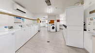 Located at 80 Forest Manor Rd in Toronto, this shared laundry facility offers a generously sized, single-floor space designed for efficient, high-capacity use. The room is bright and clean, featuring white ceramic tile flooring and white-painted walls accented by a distinctive tan horizontal tile border that runs the perimeter of the room, lending a tidy, consistent aesthetic throughout.

The facility is equipped with a substantial number of front-loading and top-loading washing machines, as well as both stacked and standalone dryers, arranged in organized rows along the walls to maximize the available floor space. Each machine is numbered for easy identification, and card-based payment terminals are conveniently positioned throughout the room for seamless transactions.

Overhead fluorescent lighting ensures the space is well-lit at all times. A folding or sorting table is available for resident use, and waste bins are placed at intervals for convenience. Informational signage and posted notices are displayed prominently on the walls, providing usage instructions and facility guidelines.

Safety is accounted for with a fire extinguisher mounted near the entrance, and a security camera is visible overhead. The layout allows for comfortable movement between machines, and a central open floor area provides ample room for residents to manage their laundry. The space is functional, orderly, and well-maintained, serving as a practical shared amenity.
