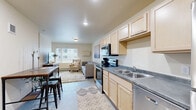 Located at 524 Center Ave in Moorhead, MN, this studio unit offers a practical and well-appointed single-floor layout. The combined kitchen and living area forms the heart of the space, designed for efficient, comfortable everyday living. The kitchen is outfitted with light maple cabinetry extending to the ceiling for generous storage, gray laminate countertops, a double stainless steel sink, and a full suite of stainless steel appliances including an electric range with over-the-range microwave, dishwasher, and refrigerator. The living area transitions seamlessly from the kitchen, featuring neutral warm-toned walls, carpeted flooring, and a large window that invites natural light and provides views of the surrounding neighborhood. A wall-mounted media console adds a functional built-in element to the living space. The bathroom features light maple cabinetry with a wide vanity, a large wall mirror, and bright overhead lighting, maintaining the consistent design palette found throughout the unit. A dedicated closet provides additional storage. The overall aesthetic is clean and modern, with a cohesive use of light wood tones, neutral finishes, and stainless steel accents throughout.