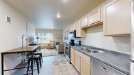 Located at 524 Center Ave in Moorhead, MN, this studio unit offers a practical and well-appointed single-floor layout. The combined kitchen and living area forms the heart of the space, designed for efficient, comfortable everyday living. The kitchen is outfitted with light maple cabinetry extending to the ceiling for generous storage, gray laminate countertops, a double stainless steel sink, and a full suite of stainless steel appliances including an electric range with over-the-range microwave, dishwasher, and refrigerator. The living area transitions seamlessly from the kitchen, featuring neutral warm-toned walls, carpeted flooring, and a large window that invites natural light and provides views of the surrounding neighborhood. A wall-mounted media console adds a functional built-in element to the living space. The bathroom features light maple cabinetry with a wide vanity, a large wall mirror, and bright overhead lighting, maintaining the consistent design palette found throughout the unit. A dedicated closet provides additional storage. The overall aesthetic is clean and modern, with a cohesive use of light wood tones, neutral finishes, and stainless steel accents throughout.