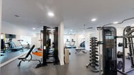Located at 514 Pleasant Dr in Durham, NC, this fitness center offers a generously sized, single-floor exercise room designed to support a full range of workout routines. The space features a clean, modern aesthetic with crisp white walls, recessed lighting, and ceiling fans throughout to ensure comfortable air circulation during workouts.

The exercise room is divided into functional zones, accommodating both cardio and strength training needs. The cardio area is equipped with treadmills, an elliptical trainer, a stationary bike, and a stair climber, each positioned for easy access and accompanied by wall-mounted televisions for entertainment. A large mirror along one wall visually expands the space and aids in form correction.

The strength training section is well-appointed with a Smith machine, a cable crossover system, a Precor functional trainer, and a dedicated dumbbell rack with a full range of free weights. Additional benches and specialty equipment round out the strength offerings. Rubber flooring throughout the entire space provides both safety and durability.

Natural light filters in through multiple windows, creating a bright and energizing atmosphere. The layout flows openly, with structural columns subtly defining training zones without impeding movement. An adjacent lounge area is visible through a passage, adding a welcoming touch to the overall facility.