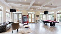 Located at 4531 Langdon Dr in Durham, NC, this single-floor community clubhouse offers a thoughtfully designed and generously proportioned gathering space. The interior is defined by light wood-look flooring, coffered and tray ceilings with recessed lighting, and neutral walls that create a bright, welcoming atmosphere throughout.

The open lounge area serves as the social heart of the space, furnished with leather seating, a live-edge wood coffee table, and cowhide ottomans arranged around a wall-mounted flat-screen TV. Large multi-pane windows and glass doors flood the area with natural light while providing direct views and access to the outdoor amenity area. A Coca-Cola vending machine adds a convenient, casual touch near the main entrance.

Adjacent to the lounge, a full-size pool table anchors the game area, offering an entertaining focal point for residents. Nearby, a well-equipped kitchen features granite countertops, gray shaker-style cabinetry, a stainless steel appliance suite including a side-by-side refrigerator and range, a subway tile backsplash, and open display shelving — ideal for community gatherings and events.

A dedicated office area with a large shared workstation, multiple computer setups, and a bold turquoise canvas artwork provides a functional and stylish workspace. Crown molding and tray ceiling details elevate this room's refined character. Together, these spaces form a versatile and polished community hub.