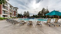 This impressive amenity space centers around a generously sized resort-style swimming pool, designed to deliver a luxurious outdoor living experience. The pool features a distinctive shallow wading area outfitted with sleek white in-water lounge chairs, ideal for relaxing while staying cool. A broad, textured concrete sun deck surrounds the pool and is lined with rows of cushioned chaise lounges, providing ample space for sunbathing and socializing. Teal market umbrellas are positioned throughout the deck, offering shaded seating options alongside bistro-height tables and chairs that create inviting spots for outdoor dining or conversation. Lush, colorful landscaping—including tropical palms, ornamental grasses, and vibrant flowering plants—frames the pool area, adding a verdant, resort-like atmosphere. A covered gazebo and additional lounge areas extend the outdoor entertaining options further around the perimeter. The adjacent indoor clubhouse lounge features a striking tiled feature wall with built-in shelving, a mounted flat-screen TV, and a fireplace, all set against neutral flooring and an open layout that transitions seamlessly to the outdoor deck through large glass doors. The multi-story brick and stone building backdrop completes the upscale, community-focused ambiance of this well-appointed amenity area.