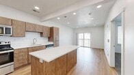Located at 61 W 400 S in Provo, Utah, this thoughtfully designed one-bedroom, one-bathroom residence offers a clean, modern aesthetic with quality finishes throughout. The open-concept kitchen and living area serves as the heart of the unit, featuring warm wood cabinetry, white quartz countertops, a central kitchen island, and a full suite of stainless steel appliances including a refrigerator, range, and microwave. A white subway tile backsplash adds a classic touch, while light hardwood flooring flows seamlessly through the main living space. Recessed lighting illuminates the area, and a sliding glass door opens to a private balcony, allowing natural light to fill the room and providing outdoor views of the surrounding neighborhood. The bedroom is carpeted for comfort, features a window with blinds for natural light, and includes access to a dedicated walk-in closet with built-in shelving and hanging rods. The full bathroom is well-appointed with hardwood-style flooring, a wood-toned vanity, and a sliding glass enclosed shower. The overall design balances contemporary style with practical functionality, creating a comfortable and cohesive living environment.