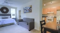 This well-appointed single-floor unit offers an open-concept layout that maximizes functionality and flow. The main living area features light-toned carpet flooring and bright white walls that lend a clean, airy feel to the space. A tufted grey sofa serves as the focal point of the living area, positioned beneath decorative wall art that adds a touch of personality to the room. Natural light filters in through a window that overlooks a lush green exterior, enhancing the sense of openness.

The sleeping area is seamlessly integrated into the living space, separated by a subtle architectural division rather than a full wall, creating a cozy yet connected environment. A dining area with a dark wood table and chair sits adjacent to the kitchen, facilitating easy transitions between meal preparation and dining.

The kitchen is compact yet thoughtfully designed, featuring light maple cabinetry, white appliances, and under-cabinet lighting that provides both practicality and warmth. Track lighting on the ceiling ensures the kitchen is well-illuminated. The overall ambiance of this unit is bright, functional, and welcoming, making efficient use of its open floor plan to create a comfortable living environment.