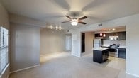 This single-floor residence offers a well-designed open-concept layout that seamlessly connects the living area to the kitchen, creating a bright and functional living environment. The expansive living space features soft, neutral-toned carpeting underfoot and a ceiling fan that anchors the room while promoting air circulation and comfort. Track lighting along one wall provides additional illumination and a modern touch to the interior.

The kitchen is a standout feature, showcasing dark espresso-toned cabinetry paired with light marble-style countertops for a striking contrast. A kitchen island offers additional prep space and casual seating potential. Stainless steel appliances, including a full-size refrigerator, contribute to the kitchen's polished, contemporary aesthetic. Warm red pendant lights hang above the island, adding a bold design accent.

The open passageway between the living and kitchen areas enhances the sense of spaciousness throughout. Neutral wall tones complement the overall palette, creating a cohesive and inviting ambiance. A hallway visible in the background hints at additional private rooms, rounding out a practical and thoughtfully arranged floor plan. The space is clean, move-in ready, and well-suited for a variety of furnishing styles.