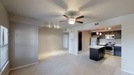 This single-floor residence offers a well-designed open-concept layout that seamlessly connects the living area to the kitchen, creating a bright and functional living environment. The expansive living space features soft, neutral-toned carpeting underfoot and a ceiling fan that anchors the room while promoting air circulation and comfort. Track lighting along one wall provides additional illumination and a modern touch to the interior.

The kitchen is a standout feature, showcasing dark espresso-toned cabinetry paired with light marble-style countertops for a striking contrast. A kitchen island offers additional prep space and casual seating potential. Stainless steel appliances, including a full-size refrigerator, contribute to the kitchen's polished, contemporary aesthetic. Warm red pendant lights hang above the island, adding a bold design accent.

The open passageway between the living and kitchen areas enhances the sense of spaciousness throughout. Neutral wall tones complement the overall palette, creating a cohesive and inviting ambiance. A hallway visible in the background hints at additional private rooms, rounding out a practical and thoughtfully arranged floor plan. The space is clean, move-in ready, and well-suited for a variety of furnishing styles.