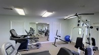 Located at 3975 I-55 N in Jackson, Mississippi, this fitness center offers a practical and functional space designed to support a well-rounded workout routine. The single-floor facility is finished with neutral-toned walls and carpeted flooring, creating a clean and comfortable environment for exercise. Bright overhead lighting ensures the space is well-illuminated throughout. A large wall mirror spans one side of the room, enhancing the sense of space and allowing users to monitor their form during workouts. The facility is equipped with a solid selection of cardio machines, including treadmills and step trainers, positioned along one side of the room for easy access. The strength training area features cable machines and weight equipment, accommodating a range of resistance exercises. Additional fitness accessories, such as an exercise ball and an ab workout bench, round out the available equipment. Windows with blinds allow for natural light while maintaining privacy. The layout is open and organized, providing ample room to move between stations comfortably. This fitness center serves as a convenient and well-equipped space for residents or members looking to maintain an active lifestyle.