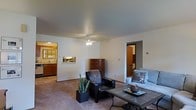 This single-floor residence offers a welcoming and functional layout designed for comfortable everyday living. The central living area is carpeted and bathed in ambient light, anchored by a ceiling fan that adds both practicality and a relaxed aesthetic. The space accommodates a seating arrangement that includes a leather armchair, a sofa with decorative pillows, and a tiled-top coffee table, creating an inviting gathering area. A wooden chest of drawers adds a warm, classic touch to the room, while a potted plant introduces a refreshing natural element. Open doorways connect the living area seamlessly to the adjoining kitchen, which features wooden cabinetry, a sink, and ample counter space, as well as to an additional room that appears to serve as a bedroom or secondary living space. The neutral wall tones and smooth ceiling throughout the home create a clean, understated backdrop that complements a variety of décor styles. The open-concept flow between rooms enhances the sense of space and connectivity, making the layout both practical and easy to navigate. Overall, the residence presents a straightforward, functional design with a warm and livable ambiance.