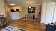 This single-floor space showcases a thoughtfully designed open-concept layout that promotes a sense of spaciousness and easy flow between living areas. Rich, wide-plank hardwood flooring runs throughout, adding warmth and character to the interior. The dining area is a focal point of the space, anchored by a sleek glass-top table surrounded by cross-back chairs, all resting on a neutral area rug. A stylish pendant light hangs above, providing both functional and ambient illumination. The walls are finished in a soft, neutral tone that complements the natural warmth of the flooring and creates a welcoming atmosphere. A bold piece of wall art depicting a longhorn steer adds a touch of personality and visual interest to the dining space. Adjacent to the dining area, the kitchen features a convenient pass-through counter with bar-height seating potential, white upper cabinetry, and dark appliances including a built-in microwave and refrigerator. The overall design balances practicality with aesthetic appeal, offering a clean, cohesive look throughout. The open sightlines and neutral palette make this a versatile and inviting space well-suited for both everyday living and entertaining.