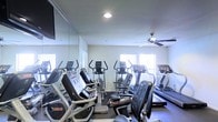 This fitness center is designed to support a well-rounded cardio-focused workout routine. The space is outfitted with a range of exercise equipment, including treadmills, elliptical trainers, upright stationary bikes, and a recumbent bike, providing users with ample options for cardiovascular training. A large floor-to-ceiling mirror runs along one wall, enhancing the sense of openness and allowing users to monitor their form during exercise. The room is illuminated by a combination of recessed ceiling lighting and natural light streaming in through multiple windows fitted with blinds, creating a bright and motivating atmosphere. A ceiling fan is mounted overhead, contributing to air circulation and overall comfort during workouts. The flooring is dark-toned and durable, appropriate for a high-traffic fitness environment. The layout is thoughtfully arranged to allow easy movement between machines. The clean, neutral-colored walls and efficient use of space give the fitness center a practical yet welcoming character, making it suitable for daily use by individuals of varying fitness levels.