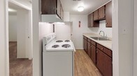 This single-floor unit offers a practical and well-organized layout designed for comfortable everyday living. The kitchen is efficiently arranged in a galley style, featuring rich dark wood cabinetry that extends from floor to ceiling, light countertops, a double-basin sink, and a freestanding electric coil range with an overhead exhaust hood. Light wood-look flooring runs throughout the kitchen, providing a clean contrast to the cabinetry. Adjacent to the kitchen, the open living area is finished with plush wall-to-wall carpeting and neutral white walls, creating a bright and versatile space. A thermostat is conveniently mounted on the wall, and a floor vent ensures climate comfort throughout. The living area connects seamlessly to a spacious bedroom, which also features carpet flooring, white walls, and a window fitted with vertical blinds that allows natural light to filter in. A wall-mounted air conditioning unit provides supplemental climate control in the bedroom. A dedicated closet with open shelving is accessible from the living space, offering generous storage. The full bathroom is finished with floor-to-ceiling white square tile surrounding a full-size soaking tub and shower combination, complemented by chrome fixtures for a clean, classic look. Overall, the space presents a straightforward and functional design throughout.