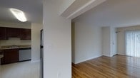 This single-floor space presents a clean, minimalist layout with a straightforward and functional design. The interior is characterized by crisp white walls and warm hardwood flooring that runs throughout the main living area, creating a cohesive and inviting atmosphere. A structural column with a beam detail subtly defines the transition between the kitchen and the open living space, adding a mild architectural element without disrupting the overall flow.

The kitchen area, partially visible to one side, features a stainless steel refrigerator and tiled flooring, contrasting with the hardwood of the adjacent living space. This distinction helps delineate the two zones while maintaining an open, connected feel. The living area is generously proportioned, offering ample room for flexible furniture arrangements.

Throughout the space, neutral tones dominate, contributing to a bright and airy ambiance. Electrical outlets are conveniently positioned along the walls. The absence of clutter and the simplicity of the finishes make this space a versatile blank canvas, well-suited to a variety of interior styles and uses.
