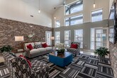 This single-story community clubhouse and leasing center is defined by its striking architectural design, combining soaring vaulted ceilings with an abundance of natural light drawn in through tall clerestory windows positioned high along the roofline. The reception area serves as the heart of the space, anchored by a large granite-topped front desk accented with pendant lighting and a bold starburst ceiling fan overhead. Stacked stone accent walls extend throughout the interior, lending a warm, textural contrast to the crisp white walls and polished concrete floors.

The lounge area is generously proportioned, offering multiple seating arrangements with neutral sofas, patterned accent chairs, and bold red throw pillows atop graphic-striped area rugs. A wall-mounted flat-screen television is integrated into one of the stacked stone feature walls, enhancing the comfortable, communal atmosphere. Large windows along the lounge's exterior wall provide views to the outdoors and flood the space with daylight.

A private office workspace is accessible from the main reception area, furnished with a traditional desk setup and accent artwork. The business center is a compact, functional room equipped with workstations, a shared printer, granite countertops, and a large-scale rose artwork as a focal point, all set atop a geometric patterned carpet.