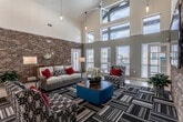 This single-story community clubhouse and leasing center is defined by its striking architectural design, combining soaring vaulted ceilings with an abundance of natural light drawn in through tall clerestory windows positioned high along the roofline. The reception area serves as the heart of the space, anchored by a large granite-topped front desk accented with pendant lighting and a bold starburst ceiling fan overhead. Stacked stone accent walls extend throughout the interior, lending a warm, textural contrast to the crisp white walls and polished concrete floors.

The lounge area is generously proportioned, offering multiple seating arrangements with neutral sofas, patterned accent chairs, and bold red throw pillows atop graphic-striped area rugs. A wall-mounted flat-screen television is integrated into one of the stacked stone feature walls, enhancing the comfortable, communal atmosphere. Large windows along the lounge's exterior wall provide views to the outdoors and flood the space with daylight.

A private office workspace is accessible from the main reception area, furnished with a traditional desk setup and accent artwork. The business center is a compact, functional room equipped with workstations, a shared printer, granite countertops, and a large-scale rose artwork as a focal point, all set atop a geometric patterned carpet.
