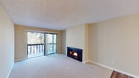 This single-floor residence offers a well-organized layout with a thoughtful mix of materials and finishes throughout. The living room serves as the heart of the home, anchored by a striking wood-burning fireplace with a dark surround and complemented by plush wall-to-wall carpeting. Large sliding glass doors open to a private balcony, flooding the space with natural light and offering views of surrounding mature trees. Adjacent to the living area, a dedicated dining space features rich hardwood-style flooring and an elegant chandelier, creating a warm and inviting atmosphere. The galley-style kitchen is efficiently designed with white cabinetry, dark countertops, stainless steel appliances including a refrigerator, microwave, and electric range, and durable hardwood-style flooring. A hallway connects the private areas of the home, where carpeted bedrooms offer a quiet retreat, each with windows that bring in natural light. Two bathrooms provide functional convenience; one features a tub and shower combination with sliding frosted glass doors, while the other includes a standalone shower with a white vanity and hardwood-style flooring. Textured ceilings are present throughout, and neutral warm-toned walls create a cohesive, move-in-ready aesthetic across the entire space.