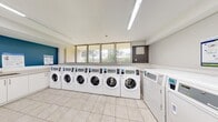 This shared laundry facility is designed with efficiency and cleanliness in mind, offering a well-organized space for residents. The room features a row of modern front-load washing machines lined along one wall beneath a series of large windows, which allow natural light to fill the space and offer views of the surrounding greenery. Opposite the washers, a matching row of top-load dryers is neatly arranged, providing ample laundry capacity. The floors are finished with light-toned ceramic tiles that contribute to the bright, hygienic atmosphere throughout. Overhead, long fluorescent light fixtures ensure consistent, even illumination across the entire space. A dedicated utility counter with white cabinetry and a sink is positioned along one wall, providing a convenient folding and preparation area. A payment kiosk and posted instructional signage are mounted nearby, streamlining the laundry process for users. Safety is addressed with a clearly visible fire extinguisher. An accent wall in bold blue adds a touch of color to the otherwise neutral palette of white and grey. A secondary entry door connects to an adjacent corridor, ensuring easy access. The overall ambiance is clean, practical, and well-maintained.