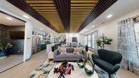 This single-floor space is thoughtfully designed with a bold, contemporary aesthetic that blends comfort and functionality across several distinct zones. The most striking architectural feature is the slatted wood ceiling treatment—alternating dark and warm-toned slats that run the length of the main gathering area, adding dimension and visual warmth overhead. Light wood-look flooring ties the open-concept layout together throughout.

The main lounge area centers around a modern sofa and accent chairs arranged on a vibrant abstract area rug, with a round white coffee table as the focal point. Adjacent to this is a secondary seating nook anchored by a textured tile wall featuring a long electric fireplace and wall-mounted television, flanked by built-in cabinetry and shelving. Large windows throughout flood the space with natural light and offer views of an outdoor area.

The kitchen stands out with deep teal cabinetry, brass hardware, white quartz countertops, open floating shelves, a geometric tile backsplash, and stainless steel appliances. A separate corridor area features starburst pendant lighting, a teal accent ceiling, flexible seating, and workstation-style tables, suggesting a co-working or business center function. The overall ambiance is energetic, colorful, and well-appointed.