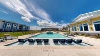 The Stella By The Bay pool area is a thoughtfully designed outdoor amenity space that delivers a resort-like experience for residents. At its center is a generously sized swimming pool with crystal-clear water, complemented by a shallow wading zone equipped with built-in in-water lounge chairs — ideal for relaxing while staying cool. A raised spa or feature pool element sits at the midpoint, adding visual interest and a layered sense of depth to the overall layout.

The expansive concrete pool deck is lined with chaise lounges and dining sets shaded by bold yellow umbrellas, creating a vibrant, coastal atmosphere under open skies. Palm trees are planted throughout the surrounding landscaped grounds, reinforcing the bayside, beach-inspired aesthetic of the community.

A covered pavilion serves as the social hub of the pool area, featuring a vaulted ceiling with ceiling fans, plush blue seating, a wall-mounted television, and an outdoor kitchen equipped with a grill — perfect for gatherings. The pavilion opens directly to the pool deck, blending indoor-outdoor living seamlessly.

The surrounding residential buildings, finished in cheerful blue and yellow coastal hues, frame the space and contribute to the cohesive, vibrant character of the Stella By The Bay community.