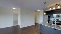 This single-floor residence offers a thoughtfully designed open-concept layout that seamlessly connects the kitchen and living areas, creating a spacious and functional flow. The kitchen is appointed with dark cabinetry, granite countertops, a stainless steel refrigerator, and a stylish pendant light fixture hanging above the counter island, adding a modern touch to the space. Rich, dark hardwood flooring runs throughout the main living area, providing a warm contrast to the crisp white walls and light ceiling. Recessed and flush-mount ceiling lighting ensures the space remains well-lit throughout. The living area offers generous wall space, ideal for flexible furniture arrangements, and connects to an adjacent room with carpeted flooring — providing a softer, quieter retreat. A hallway visible in the background leads to additional rooms and closet space, enhancing the overall practicality of the layout. The clean, neutral color palette and modern finishes throughout give the space a contemporary, move-in-ready feel that balances both style and comfort.