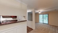 This single-floor residence showcases a thoughtfully designed open layout that connects the kitchen and living areas in a cohesive, functional flow. The kitchen features rich dark granite countertops that contrast beautifully against crisp white cabinetry, including raised-panel lower cabinets and upper storage units with a classic tile backsplash. A decorative pass-through opening, framed by white columns and crown molding, bridges the kitchen and living space, adding architectural character while maintaining an open, airy feel.

The transition from kitchen to living area is marked by a shift from hardwood-style flooring to plush carpeting, delineating the spaces naturally. Crown molding is carried throughout, lending a refined, polished look to the interior. A ceiling fan with a light fixture is centrally positioned in the living area, providing both comfort and ambient lighting.

Large vertical blinds frame a full-length sliding glass door that draws in generous natural light and offers views of lush greenery outside, enhancing the overall brightness and welcoming ambiance of the space.