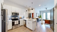 This thoughtfully designed 1-bedroom, 2-bathroom residence offers a single-floor open-concept layout that maximizes every inch of its compact footprint. Light wood-tone flooring flows throughout the main living areas, connecting the kitchen, dining, and living spaces in a seamless continuum. The modern kitchen is a standout feature, equipped with stainless steel appliances including a range, microwave, and dishwasher, complemented by crisp white shaker cabinetry, a blue tile backsplash, quartz countertops, and a central island with a built-in sink and pendant lighting overhead. The living room is bright and welcoming, anchored by large floor-to-ceiling glass doors that open to a private balcony, allowing abundant natural light and outdoor views. The bedroom offers a cozy retreat with plush carpeting, a generous window with outdoor views, and direct access to a walk-in closet. The primary bathroom features a soaking tub and shower combination with marble-style tile surround, a granite-topped vanity, and wood-look flooring. A dedicated in-unit laundry area with a full-size washer and dryer adds everyday convenience. The overall ambiance is modern and inviting, with clean lines and contemporary finishes throughout.
