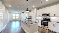 This well-appointed single-floor residence offers 2 bedrooms, 2 bathrooms, and 2 closets in a thoughtfully designed open layout. The kitchen and living area share a seamless open-concept space anchored by a large central island with dark granite countertops and an undermount sink, illuminated by stylish bronze pendant lights. White shaker-style cabinetry, a classic subway tile backsplash, and stainless steel appliances — including a range, microwave, and refrigerator — complete the kitchen. Rich hardwood flooring flows throughout the main living areas, complemented by neutral-toned walls and recessed lighting that create a bright, clean atmosphere. Natural light pours in through multiple windows. Both bedrooms feature plush carpet flooring and ceiling fans, with windows offering outdoor views. The primary bedroom is generously sized and connects to an en suite bathroom showcasing a double-sink vanity with dark granite countertops, backlit mirrors, white cabinetry with matte black hardware, hardwood floors, and a glass-enclosed shower. A well-organized walk-in closet with built-in shelving and hanging rods adds valuable storage. The second bathroom mirrors the same upscale finishes. The home's cohesive modern aesthetic, quality materials, and functional design make it a refined and comfortable living space.