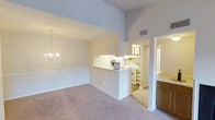 This single-floor residence showcases a thoughtfully designed open-concept layout that seamlessly connects the living, dining, and kitchen areas. The dining space immediately draws the eye upward with its impressively vaulted ceilings, which lend an expansive, airy quality to the interior. An elegant chandelier anchors the dining area, adding a refined touch to the otherwise clean and neutral palette. Soft, wall-to-wall carpeting in warm beige tones runs through the main living spaces, contributing to a comfortable and inviting atmosphere. A half-wall pass-through connects the main living area to the kitchen, enhancing the open feel while maintaining a subtle sense of separation. The kitchen is well-appointed with light cabinetry, a white appliance suite including a range and refrigerator, and a bright, functional layout. A second cabinetry area with warm wood-toned lower cabinets and light countertops is visible adjacent to the main space, offering additional storage and prep area. The walls throughout are finished in soft, neutral tones that complement the natural light filtering into the space. HVAC vents are positioned at ceiling level, ensuring efficient climate control. Overall, the space presents a bright, clean, and functional interior well-suited for comfortable everyday living.