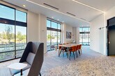 This contemporary single-floor communal space is thoughtfully designed to accommodate both collaborative and relaxed interactions. The open-plan layout seamlessly connects a living area and a dining area, unified by soft, patterned carpeting and crisp white walls that contribute to a clean, modern aesthetic.

Floor-to-ceiling windows line multiple walls throughout the space, flooding the interior with abundant natural light and offering expansive views of the surrounding urban streetscape. Linear LED lighting integrated into the ceiling complements the natural light, maintaining a bright and welcoming atmosphere throughout the day.

The living area features high-back upholstered lounge chairs in cool grey tones, arranged for intimate conversation alongside small side tables. The dining area is anchored by a rectangular wood-topped table surrounded by sleek amber-orange leather chairs, providing a functional yet stylish setting for group gatherings or meetings.

A separate, enclosed meeting room is also available, furnished with a compact wooden conference table, ergonomic chairs, and a wall-mounted display screen, making it well-suited for focused discussions or presentations. Additional amenities visible in the space include a printer station and small café-style seating nooks near the windows.

Bold, colorful artwork adorns the walls, adding artistic character and warmth to the otherwise neutral palette. A deep blue accent wall with glass double doors serves as a striking architectural focal point, enhancing the overall design cohesion of this versatile, professionally appointed space.