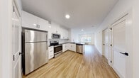 Located at 27 Colfax Ave in Pompton Lakes, NJ, this thoughtfully designed single-floor unit offers a clean, modern aesthetic with an open-concept layout that seamlessly connects the kitchen and living areas. The kitchen is a standout feature, equipped with a full suite of stainless steel appliances including a refrigerator, range, microwave, and dishwasher. White shaker-style cabinetry reaches toward the ceiling, complemented by a classic subway tile backsplash and sleek dark countertops that provide striking contrast. A peninsula with additional counter space enhances both functionality and flow. Light wood-tone vinyl flooring runs consistently throughout the unit, contributing to a bright, cohesive atmosphere. The living area benefits from a large picture window that invites natural light and frames views of the surrounding streetscape. The bedroom spaces feature crisp white walls and the same warm flooring, offering a versatile, move-in-ready canvas. The bathroom is finished with large-format light gray wall tiles surrounding a soaking tub and shower combination, paired with chrome fixtures for a polished look. Recessed lighting throughout the unit ensures even, modern illumination. The entry area provides practical access with a utility closet nearby, adding to the overall functionality of this well-appointed home.