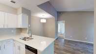 This single-story unit showcases a thoughtfully designed open-concept floor plan with neutral gray tones and wood-look flooring throughout the main living areas. The living room is anchored by a tile-surround fireplace and benefits from soaring vaulted ceilings that enhance the sense of space. A dedicated entryway provides a welcoming transition into the home.

The kitchen is well-appointed with white shaker-style cabinetry, white countertops, a light subway tile backsplash, and stainless steel appliances including an electric range and over-the-range microwave. A peninsula with an integrated sink and dishwasher connects the kitchen to the adjacent dining area, which is illuminated by a pendant light fixture.

Bedrooms feature plush carpeting, ceiling fans, and windows with blinds that allow for natural light and outdoor views. At least one bedroom includes direct access to a bathroom, which offers a white vanity, large mirror, and a tiled tub-shower combination with wood-look flooring. A second bathroom follows a similar clean, functional design.

A dedicated laundry area houses a full-size washer and dryer. Textured ceilings are present throughout, completing the practical and comfortable character of this single-story residence.