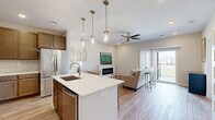 Located on Legends Drive in Champaign, Illinois, this single-floor residence offers a thoughtfully designed open-concept layout that seamlessly connects the kitchen, dining, and living areas. The kitchen is a standout feature, equipped with rich warm-toned cabinetry, stainless steel appliances including a refrigerator, range, and microwave, a classic subway tile backsplash, and a large central island with a white quartz countertop, undermount sink, and pendant lighting overhead. Light wood-style flooring runs throughout the main living areas, contributing to a bright and cohesive aesthetic. The living room centers around a sleek linear fireplace and opens directly to a private balcony through a sliding glass door, offering pleasant views of the surrounding neighborhood. A ceiling fan adds comfort to both the living room and bedroom areas. The bedrooms feature plush carpeting and natural light from well-placed windows. The bathroom includes a warm wood vanity with white quartz countertop and contemporary fixtures. A dedicated in-unit laundry area with a washer and additional hallway storage provide practical convenience. Neutral tones and clean lines throughout create a calm, modern ambiance well-suited for everyday living.