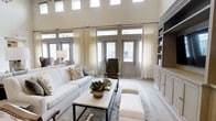 This single-story space showcases an impressive great room defined by dramatic double-height ceilings that create an expansive, airy atmosphere. A wall of French doors, flanked by large transom windows above, floods the interior with natural light while offering a seamless visual connection to the outdoors. Sheer floor-to-ceiling drapery panels frame the doors elegantly, softening the architectural lines and adding a refined, layered texture to the space.

The living area is thoughtfully arranged with a neutral-toned sofa, upholstered seating, and a rectangular coffee table anchored by a decorative botanical centerpiece. Light hardwood flooring runs throughout, complementing the warm yet muted color palette of whites, grays, and soft taupes that define the interior aesthetic.

A substantial built-in shelving and cabinetry unit lines one wall, combining open display shelves with closed lower cabinets finished in a contrasting wood tone, providing both functional storage and a striking design focal point. The overall ambiance is transitional — blending classic architectural details with clean, contemporary furnishings for a polished and welcoming environment.