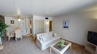 This single-floor unit is laid out to offer a functional and cohesive living environment. The living room serves as the central gathering space, finished with wall-to-wall carpet in a neutral tone and crisp white walls. A sliding glass door with vertical blinds fills the room with natural light and provides direct access to an outdoor patio area. A glass-top coffee table and a striped sofa complement the light, airy aesthetic, while a framed artwork piece and louvered closet doors add modest architectural character.

The galley-style kitchen is efficiently designed, with warm oak cabinetry lining both sides and ample counter space for meal preparation. White appliances, including a refrigerator, electric range, and built-in microwave, are neatly integrated. A skylight-style ceiling fixture ensures the kitchen remains well-lit throughout the day.

The bedroom is a comfortable, carpeted room with a natural window that allows in soft light. A multi-drawer dresser with a round mirror provides practical storage, and additional drawer space is available via a tall chest unit.

The bathroom features a full tub and shower combination with clean white surroundings and tile flooring, completing this straightforward and well-maintained unit.